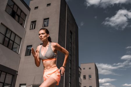Young. serious sporty brunette woman running past buildings wearing shorts and a bikini bra.