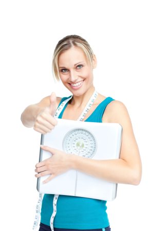 How to make fitness a habit - smiling, blonde woman with a measuring tape giving a thumbs up while holding a weight scale